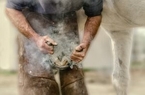 A beautifully processed photo of a farrier fitting a new shoe on a horse with the ssmoke from the hot shoe being burnt into the nail billowing around.