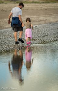 A photo of a man and his daughter reflected in a puddle as they stroll along the beach.