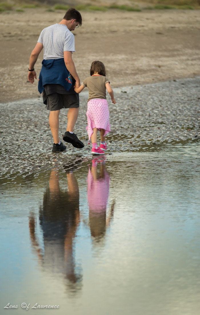 A photo of a man and his daughter reflected in a puddle as they stroll along the beach.