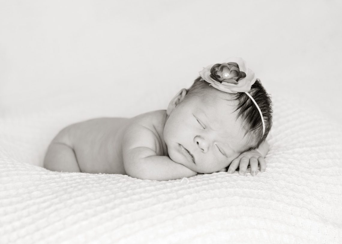 A beautiful black and white photo of a nude Newborn girl with only a simple headpiece prop lying asleep on a gorgeous white textured wrap