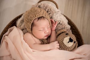 A photo of a gorgeous Newborn by photographed with props consisting of a round basket, flokati throw, brown Teddy Bear and beige wrap and matching cap
